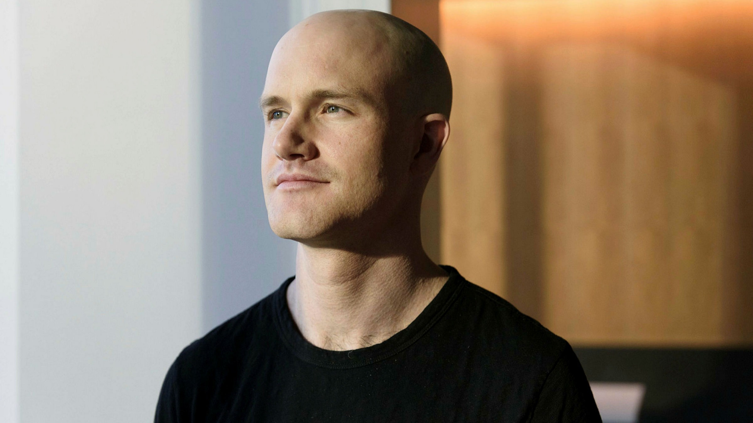 A person in a black shirt looks contemplatively off-camera in a softly lit room with a wooden backdrop.
