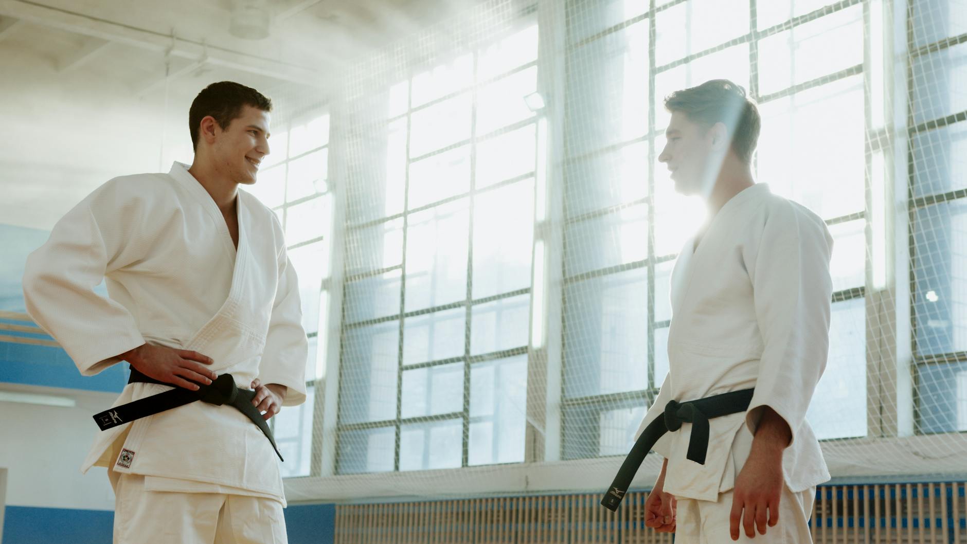 Two martial artists in white uniforms, smiling during a training session in a sunlit dojo.